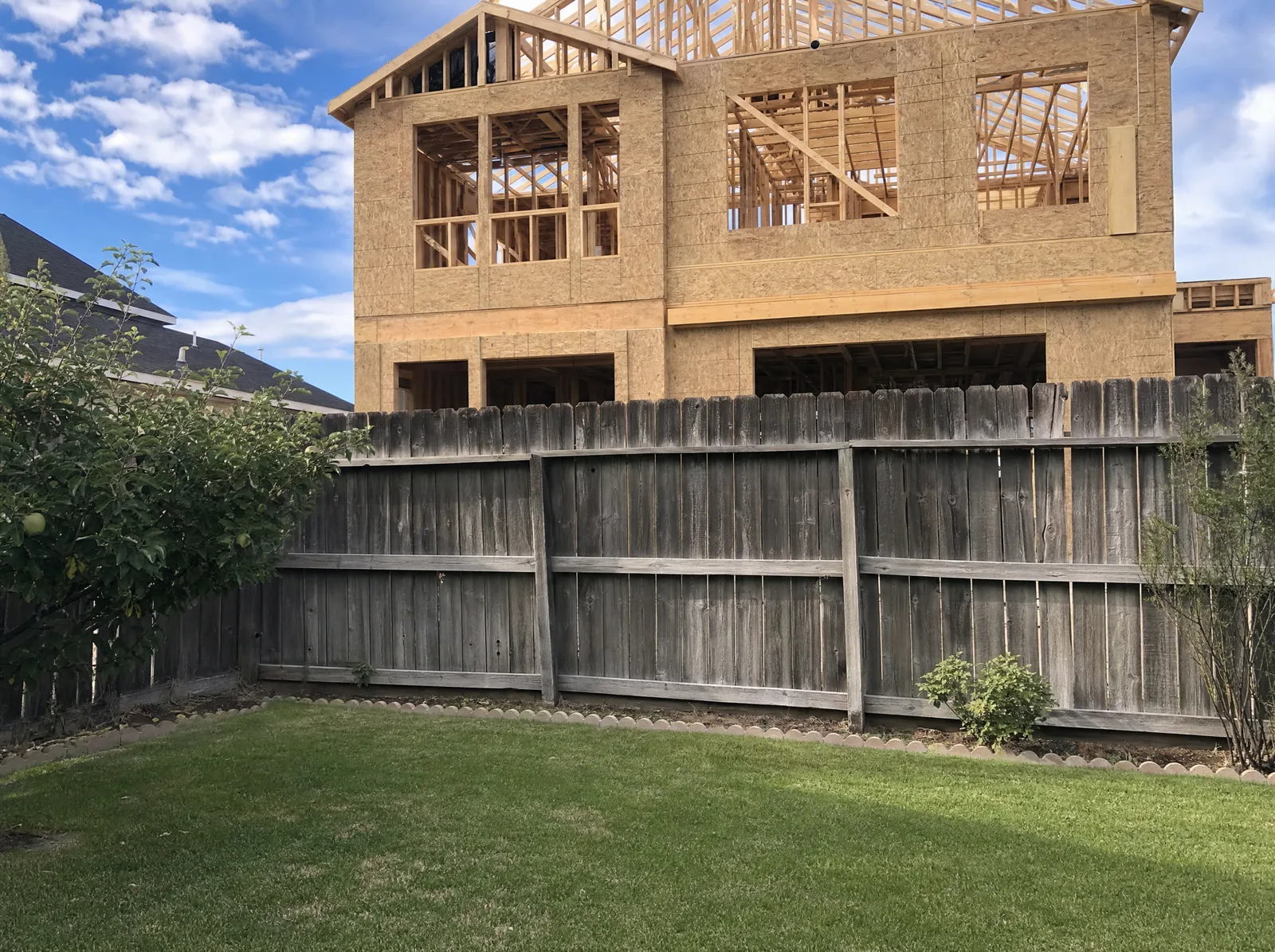 Older wood privacy fence along a back property line with a tall two-story house under construction looming directly behind it